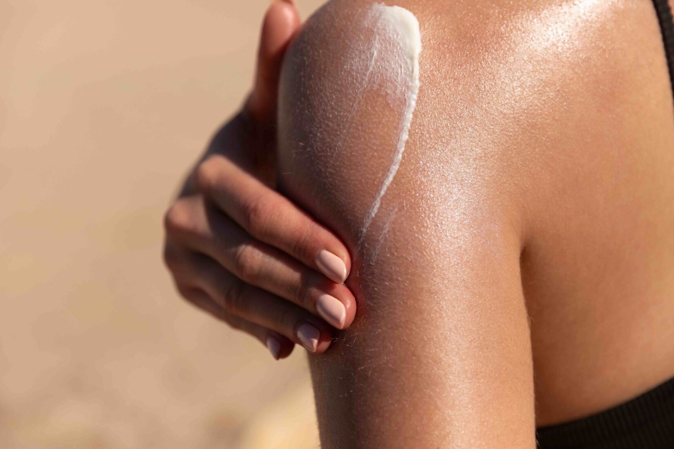 Young woman applying sun cream or sunscreen on her tanned shoulder to protect her skin from the sun. Shot on a sunny day with blurry sand in the background
